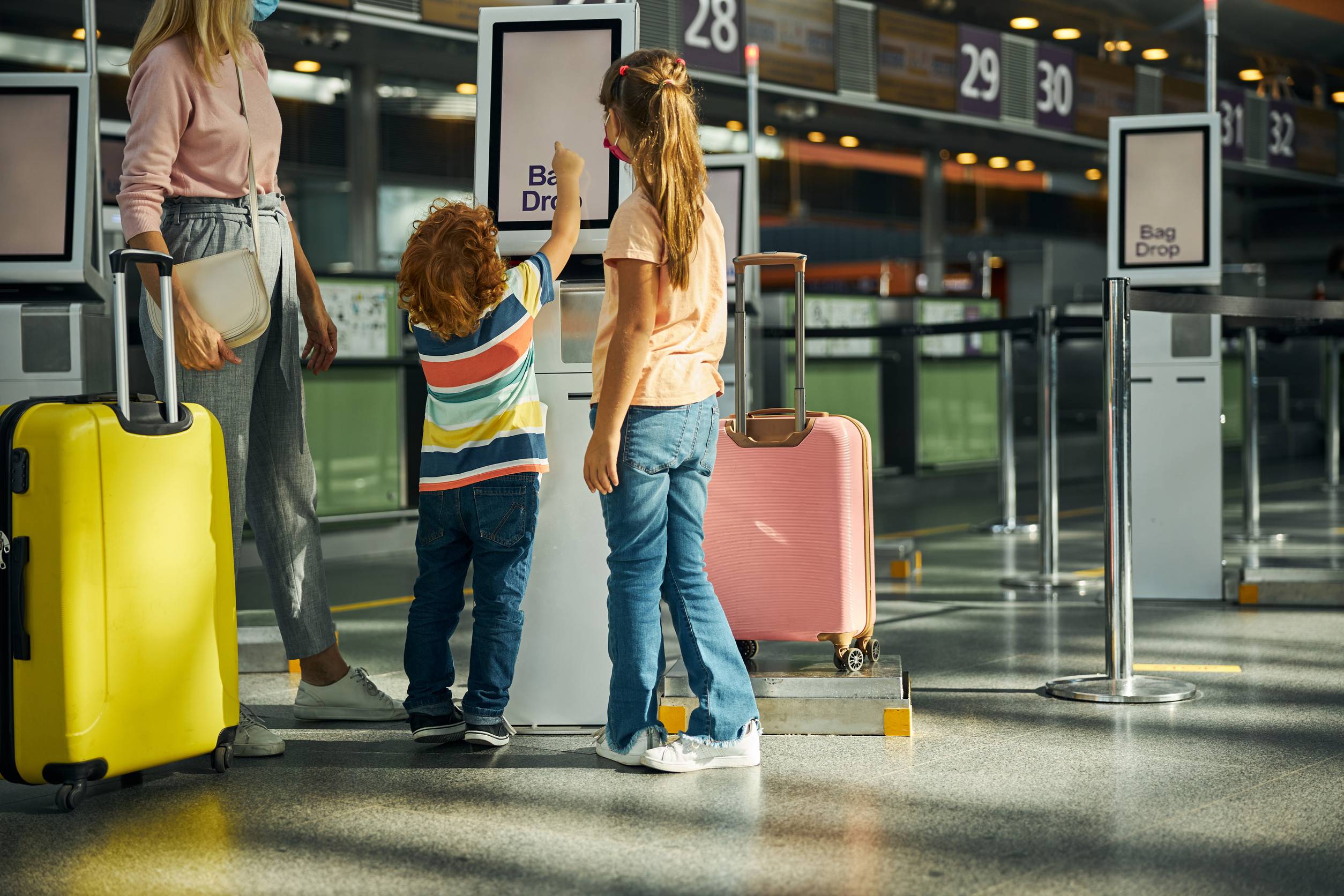 Interested Kid Raising His Hand And Tapping On A Screen Of A Baggage Drop Terminal With His Family